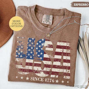 May include: Espresso-colored t-shirt with a distressed USA flag design. The shirt features the text "USA" in red, white, and blue, with "SINCE 1776" below. A silver necklace with a pendant rests on the shirt.