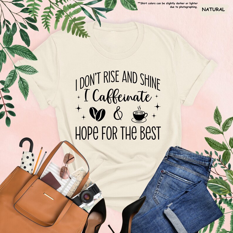 May include: Cream-colored t-shirt with the text "I Don't Rise and Shine, I Caffeinate, Hope for the Best" in black. The design includes coffee beans and a coffee cup. The shirt is paired with blue jeans and accessories.