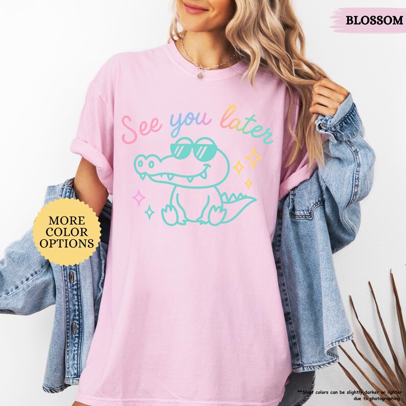 Puede incluir: Camiseta rosa claro con un cocodrilo de dibujos animados con gafas de sol y el texto "See you later" en colores pastel. La camiseta tambi&eacute;n tiene el texto "More Color Options" y "Blossom".