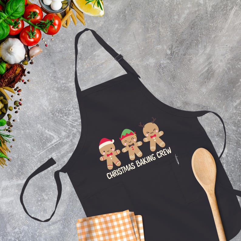 Op de afbeelding: Zwarte schort met een witte afbeelding van drie gingerbreadmen die kerstpetten dragen en de tekst "Christmas Baking Crew".
