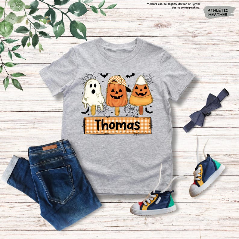 Może przedstawiać: Szary melanżowy t-shirt z kr&oacute;tkim rękawem z grafiką przedstawiającą trzy lody na patyku w stylu Halloween: duszka, dynię w czapce oraz cukierek kukurydziany. Poniżej znajduje się pomarańczowy prostokąt w kratkę z nadrukowanym imieniem Thomas.