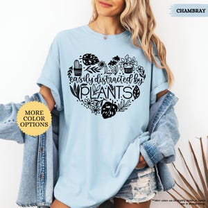 Pu&ograve; includere: Una maglietta blu chiaro con un design a forma di cuore nero che presenta il testo "easily distracted by PLANTS" e varie illustrazioni di piante.