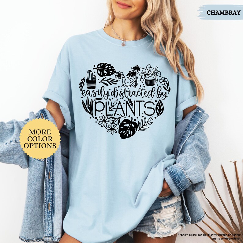 Pu&ograve; includere: Una maglietta blu chiaro con un design a forma di cuore nero che presenta il testo "easily distracted by PLANTS" e varie illustrazioni di piante.