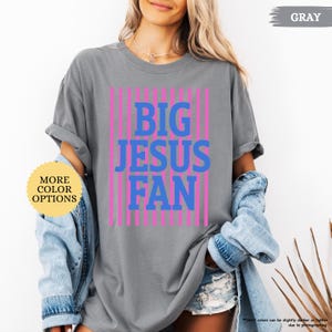 May include: Gray t-shirt with the text "BIG JESUS FAN" in blue and pink vertical stripes. The shirt has rolled-up sleeves and is paired with a denim jacket. A button on the left says "MORE COLOR OPTIONS".