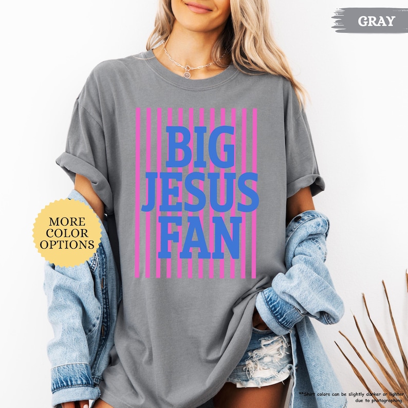 May include: Gray t-shirt with the text "BIG JESUS FAN" in blue and pink vertical stripes. The shirt has rolled-up sleeves and is paired with a denim jacket. A button on the left says "MORE COLOR OPTIONS".