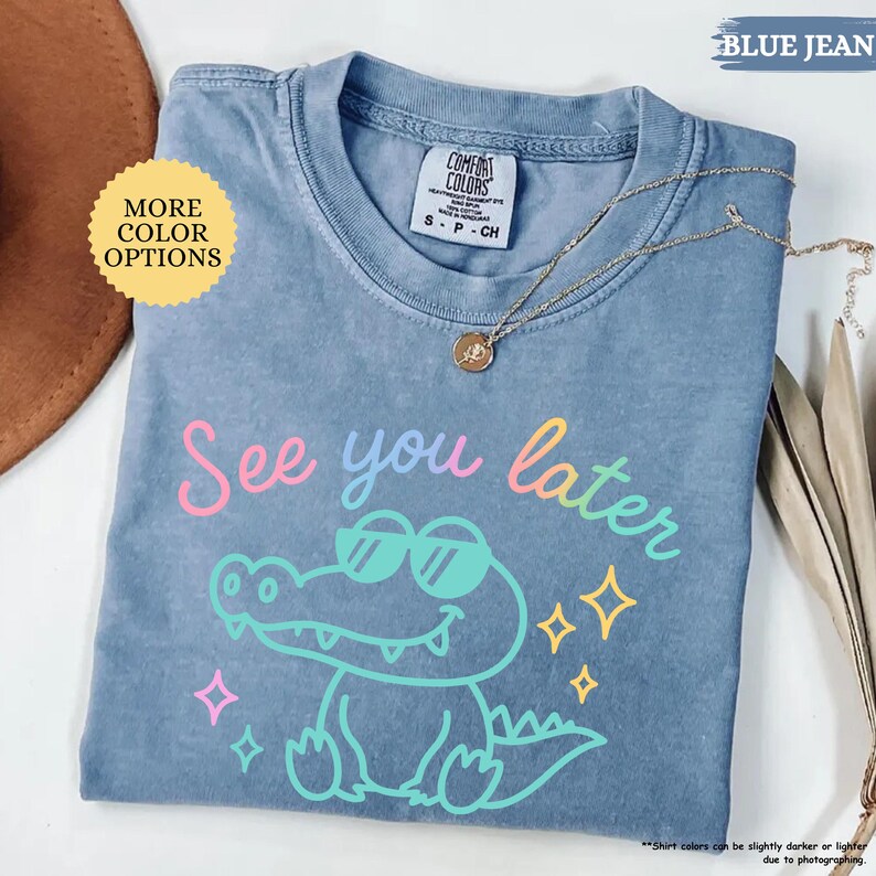 Puede incluir: Camiseta azul vaquero con la frase "See you later" y un cocodrilo de dibujos animados con gafas de sol. La camiseta tiene cuello redondo y mangas cortas. Un collar dorado con un colgante est&aacute; drapeado sobre la camiseta.