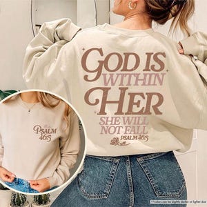 Puede incluir: Una sudadera de color arena con el texto "GOD IS WITHIN HER SHE WILL NOT FALL PSALM 46:5" en letras marrones y rosas. La sudadera tiene cuello redondo y mangas largas. Una imagen m&aacute;s peque&ntilde;a de la sudadera se muestra en un recuadro circular.