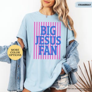 May include: Light blue t-shirt with the text "BIG JESUS FAN" in blue letters, set against a pink striped background. The shirt is a casual style, with rolled-up sleeves. The word "CHAMBRAY" is written in the top right corner.