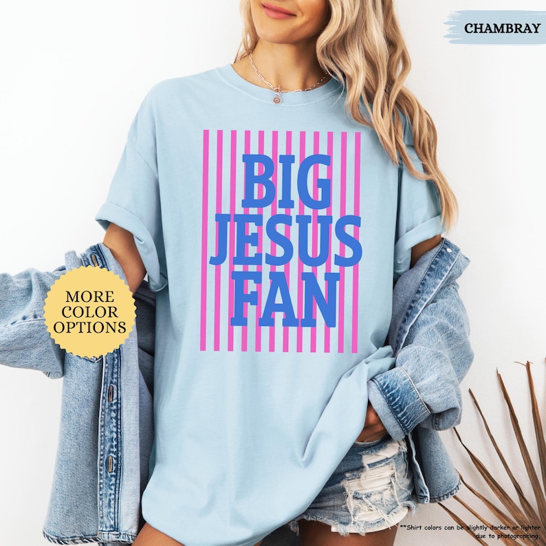 May include: Light blue t-shirt with the text "BIG JESUS FAN" in blue letters, set against a pink striped background. The shirt is a casual style, with rolled-up sleeves. The word "CHAMBRAY" is written in the top right corner.