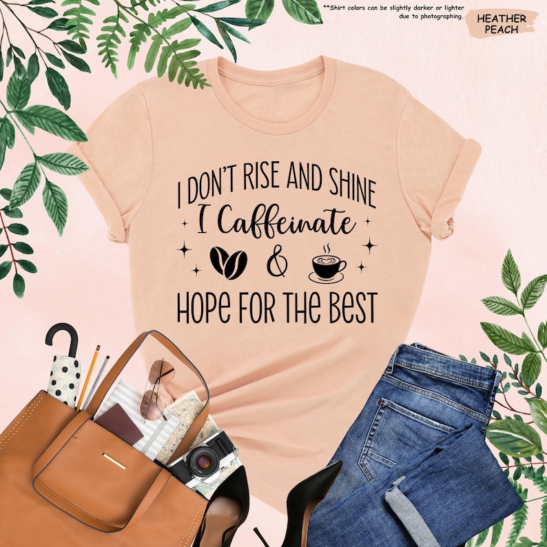 May include: A peach-colored t-shirt with the text "I DON'T RISE AND SHINE I Caffeinate & HOPE FOR THE BEST" in black. The design includes coffee beans and a coffee cup. The shirt is paired with blue jeans, a brown purse, and black heels.