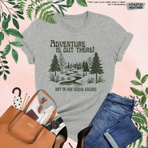 May include: Athletic heather gray t-shirt with the text "Adventure is Out There!" and "But So Are Serial Killers" in a dark green font. The graphic features a forest scene with a winding river. The shirt is paired with blue jeans.