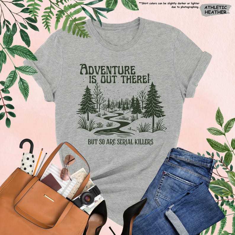 May include: Athletic heather gray t-shirt with the text "Adventure is Out There!" and "But So Are Serial Killers" in a dark green font. The graphic features a forest scene with a winding river. The shirt is paired with blue jeans.