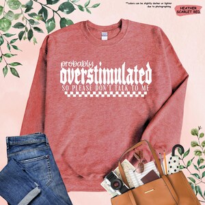 May include: A heather scarlet red sweatshirt with the text "probably overstimulated so please don't talk to me" in white. The text is in a gothic font and is surrounded by a checkered pattern.