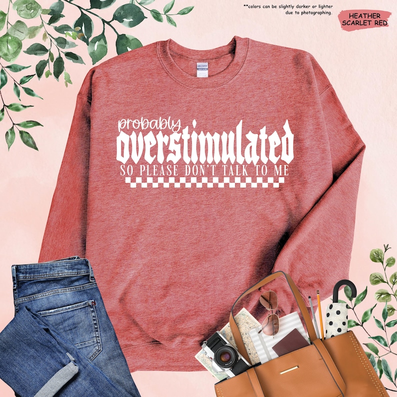 May include: A heather scarlet red sweatshirt with the text "probably overstimulated so please don't talk to me" in white. The text is in a gothic font and is surrounded by a checkered pattern.