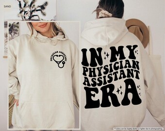 May include: A cream-colored hoodie with a black graphic that says "In My Physician Assistant Era" with stars and a stethoscope graphic.