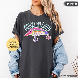 以下が含まれることがあります： チャコールグレーの半袖Tシャツ。ライトブルーの文字で「CATCH YA LATER」と書かれ、その下にカラフルな釣り用ルアーのグラフィックが描かれています。ルアーにはピンク、オレンジ、ブルーのドットや星の模様が施され、2つのフックが付いています。