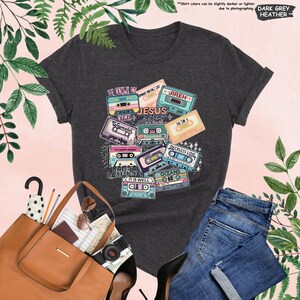 May include: Dark grey heather t-shirt featuring a colorful graphic of cassette tapes with religious phrases like "Jesus", "Waymaker", and "Reckless Love". The shirt is paired with blue jeans and a brown tote bag.