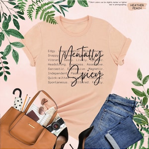 K&ouml;nnte beinhalten: Ein pfirsichfarbenes T-Shirt mit den Worten "Mentally Spicy" und beschreibenden Adjektiven. Das Shirt wird mit einer blauen Jeans, einer braunen Tragetasche, schwarzen Abs&auml;tzen und Accessoires kombiniert. Der Text auf dem Shirt lautet: Edgy, Unapologetic, Mischievous, Snappy, Clever, Sassy, Lively, Dynamic.