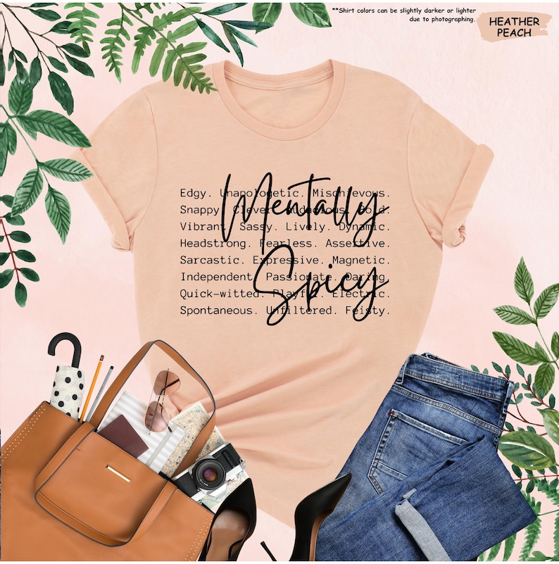 K&ouml;nnte beinhalten: Ein pfirsichfarbenes T-Shirt mit den Worten "Mentally Spicy" und beschreibenden Adjektiven. Das Shirt wird mit einer blauen Jeans, einer braunen Tragetasche, schwarzen Abs&auml;tzen und Accessoires kombiniert. Der Text auf dem Shirt lautet: Edgy, Unapologetic, Mischievous, Snappy, Clever, Sassy, Lively, Dynamic.