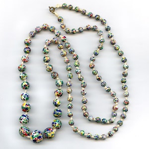 May include: A long necklace with colorful, speckled beads in shades of blue, green, yellow, and red. The beads are strung on a cream-colored cord, and the necklace has a gold-colored clasp. The necklace is likely vintage or handmade.