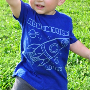 Peut inclure: Un jeune enfant portant un t-shirt bleu avec un motif de fusée et le mot "Adventure". L'enfant est sur de l'herbe verte, avec un mur de briques en arrière-plan.
