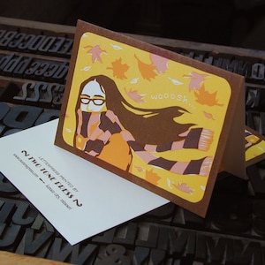 May include: A letterpress printed card with a brown background and a yellow, pink, and brown illustration of a person with long brown hair wearing a scarf and walking in the wind. The text "wooosh..." is written in white above the person. The card also includes the text "LETTERPRESS PRINTED BY TWO TONE PRESS" and "kansas city, missouri".
