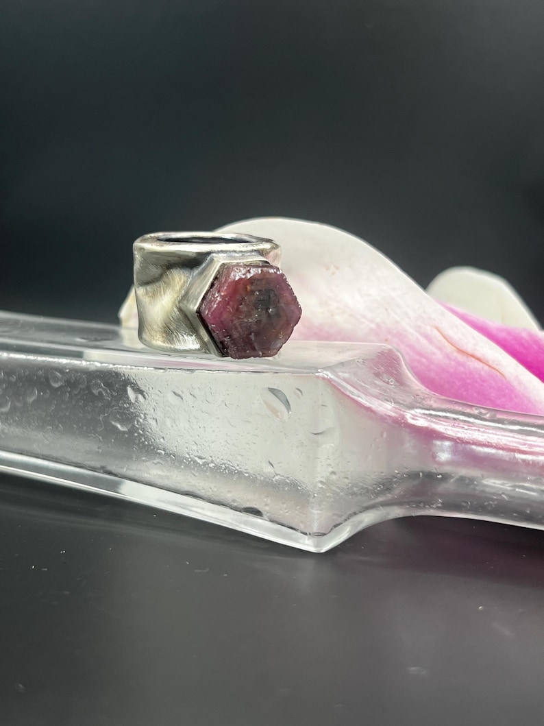 May include: A silver ring with a large, raw ruby gemstone. The ring has a thick band and a geometric setting that holds the ruby. The ruby is a deep red color and has a rough, natural texture. The ring is displayed on a clear glass surface.
