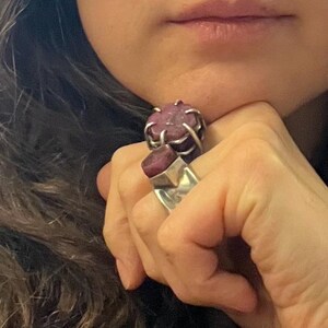 May include: Two silver rings with large, raw ruby stones. One ring features a cage-like setting around the ruby, while the other has a rectangular stone set in a simple band. The rings are worn on a hand.