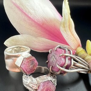May include: Three silver rings with large, raw, pink gemstones are displayed against a backdrop of a white and pink magnolia flower. The rings have unique, textured settings, showcasing the natural beauty of the stones.