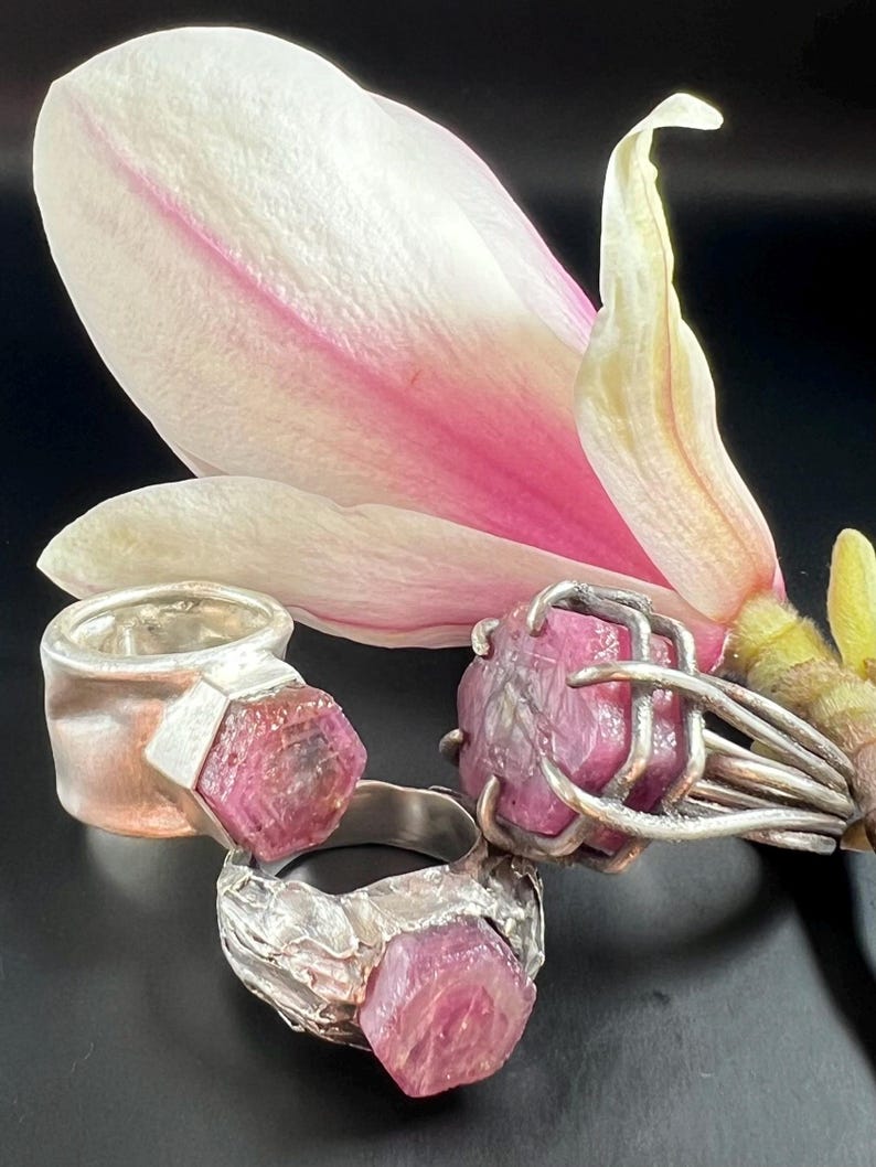 May include: Three silver rings with large, raw, pink gemstones are displayed against a backdrop of a white and pink magnolia flower. The rings have unique, textured settings, showcasing the natural beauty of the stones.