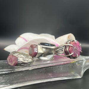 May include: Three silver rings with raw ruby gemstones. The rings are displayed on a clear glass surface with a white and pink flower in the background. The rubies are a deep pink color.