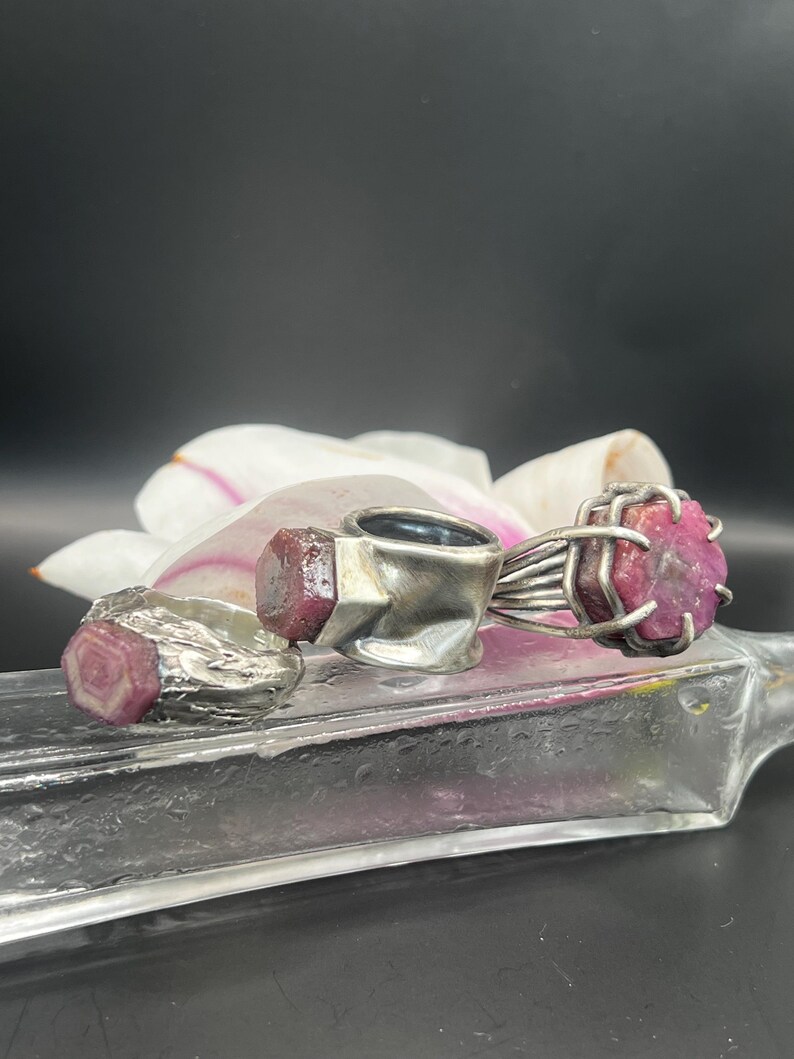 May include: Three silver rings with raw ruby gemstones. The rings are displayed on a clear glass surface with a white and pink flower in the background. The rubies are a deep pink color.