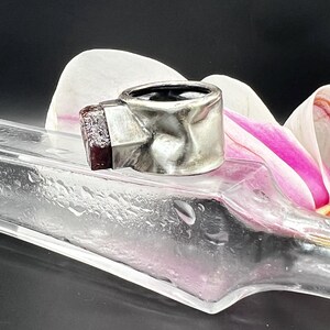 May include: A silver ring with a textured band and a rectangular, dark red gemstone. The ring is set against a blurred background with a clear glass object and a white and pink flower.
