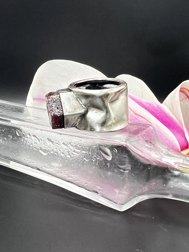 May include: A silver ring with a textured band and a rectangular, dark red gemstone. The ring is set against a blurred background with a clear glass object and a white and pink flower.