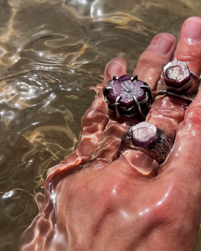 May include: Close-up of a hand holding four gemstone rings in water. The rings feature raw, uncut purple and pink gemstones set in silver-toned metal. The water reflects light, creating a shimmering effect.