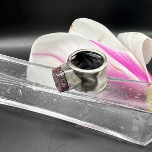 May include: A silver ring with a dark interior and a rectangular, textured, reddish-brown stone. The ring is set against a backdrop of a white and pink flower and a clear glass tray.