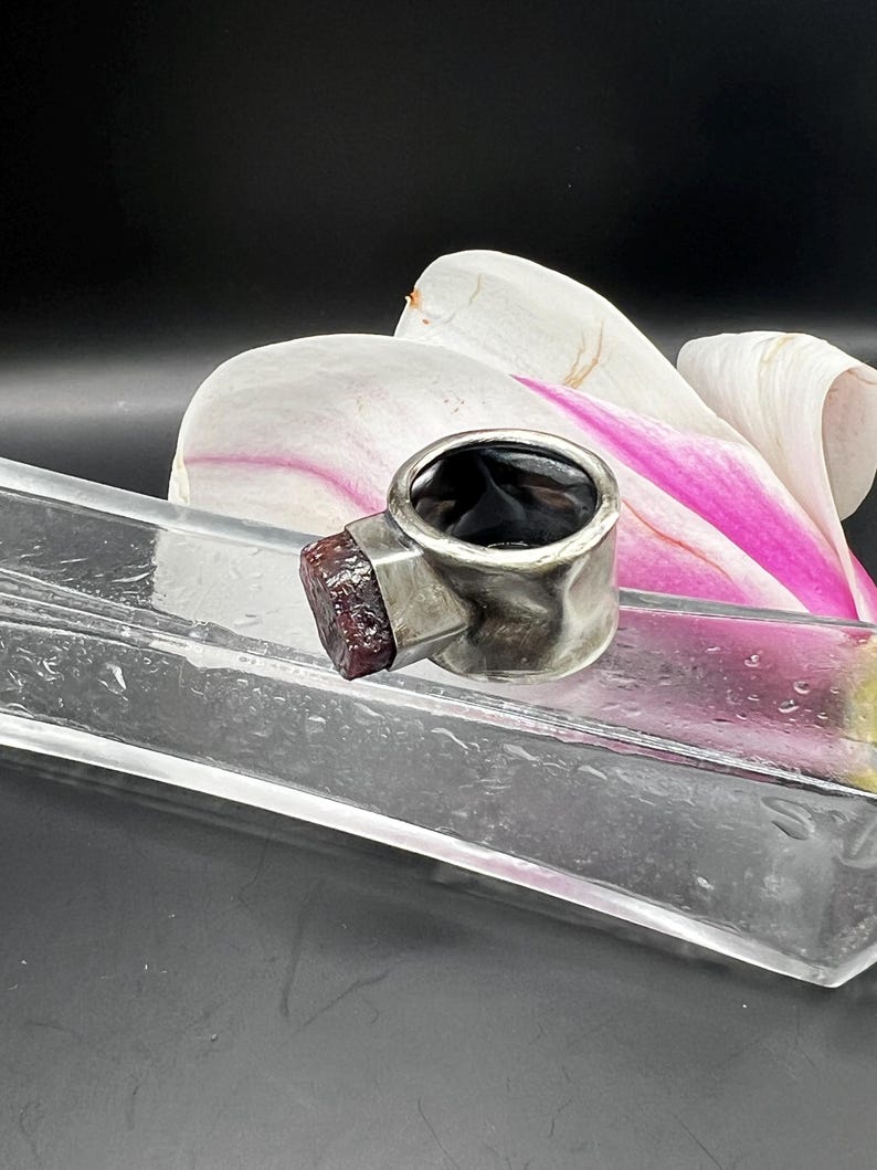 May include: A silver ring with a dark interior and a rectangular, textured, reddish-brown stone. The ring is set against a backdrop of a white and pink flower and a clear glass tray.