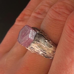 Könnte beinhalten: Ein Silberring mit einem großen, facettierten, rosa Edelstein. Der Ring hat ein strukturiertes, organisches Design um den Edelstein herum, was eine einzigartige und natürliche Ästhetik schafft. Der Edelstein hat eine lebendige rosa Farbe.