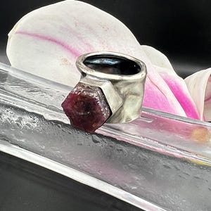 May include: A silver ring with a raw, dark red gemstone. The ring has a thick band with a textured finish. The gemstone is a deep burgundy color and is set in a geometric design. The ring is displayed on a clear surface with a white and pink flower in the background.
