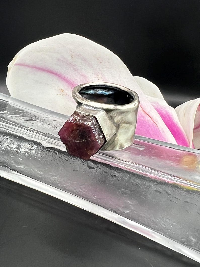 May include: A silver ring with a raw, dark red gemstone. The ring has a thick band with a textured finish. The gemstone is a deep burgundy color and is set in a geometric design. The ring is displayed on a clear surface with a white and pink flower in the background.