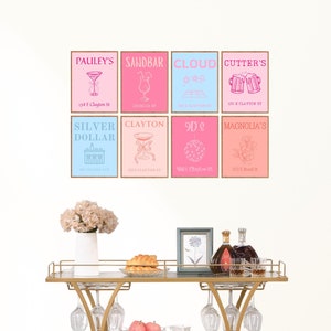 May include: A gold metal bar cart with three glass shelves. The cart has a wine rack on the bottom shelf and a glass top shelf. The cart is decorated with various signs with pink, blue, and white backgrounds. The signs have names of bars and restaurants with addresses.