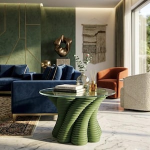 May include: A contemporary living room with a round glass-topped coffee table with a green, sculptural base. The room features a blue velvet sofa, an orange armchair, and a patterned rug. The walls are green and white, with a large window.