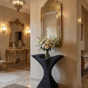 Pode incluir: Uma mesa consola preta com um tampo curvo e em camadas e uma base cónica, com um vaso de vidro transparente contendo um ramo de rosas brancas e creme, sob um espelho alto e arqueado de metal dourado com gravações intrincadas.