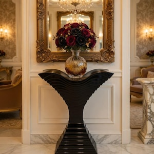 May include: A decorative black console table with a unique wave-like design supporting a glass vase filled with red and black roses. The table is in front of a gold-framed mirror, reflecting a chandelier.
