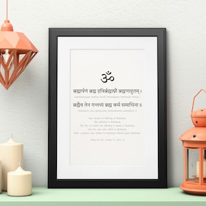 Puede incluir: Una impresión enmarcada en negro con fondo blanco y texto negro en sánscrito e inglés. El texto incluye el símbolo sánscrito Om y la frase "Brahmarpanam Brahma Havirbrahmaagnau Brahmanaahutam | Brahmauva Tena Gantavya Brahma Karma Samadhina ||". La impresión está sobre una superficie blanca con una lámpara naranja claro y dos velas blancas.