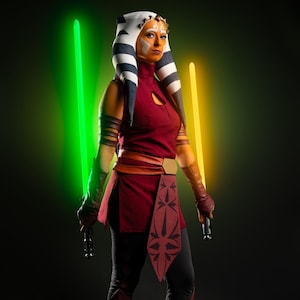 Peut inclure: Une femme vêtue d'un costume de Ahsoka Tano, un personnage de Star Wars, avec une tenue rouge et noire, une coiffe blanche et noire, et deux sabres laser, l'un vert et l'autre jaune, devant un fond noir.
