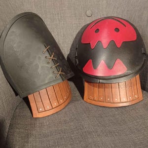 Puede incluir: Dos protectores de brazo de cuero negro con ribetes de cuero marrón y un diseño de cara de monstruo rojo. Los protectores de brazo están sobre una superficie de tela gris.