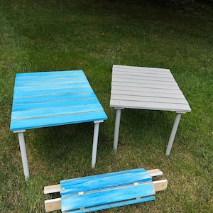 May include: Two unfinished wooden tables, one painted light blue and the other grey, with a third blue table top lying on the grass.