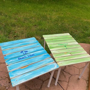 Peut inclure: Deux petites tables en bois avec des pieds blancs. La table de gauche est peinte en bleu et dit "Beach Vibes" avec un graphique de palmier. La table de droite est peinte en vert et dit "Happy Hour" avec un graphique de verre à cocktail.
