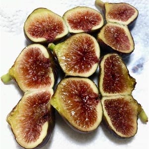Peut inclure: Une sélection de figues fraîches, coupées en deux pour révéler leur intérieur rouge vif. Les figues ont une peau verte et jaune, avec une base violet foncé. L'arrangement est sur une surface blanche, mettant en valeur les couleurs et les textures naturelles du fruit.